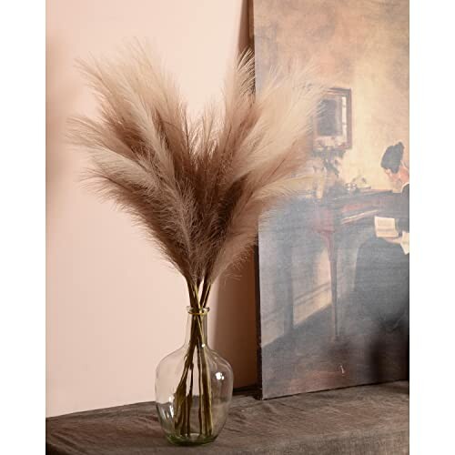 Vase with pampas grass beside a painting on a table.